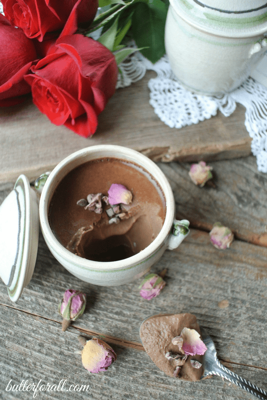 A petit chocolate rose pot de crème topped with rose petals on a wood table with red roses.